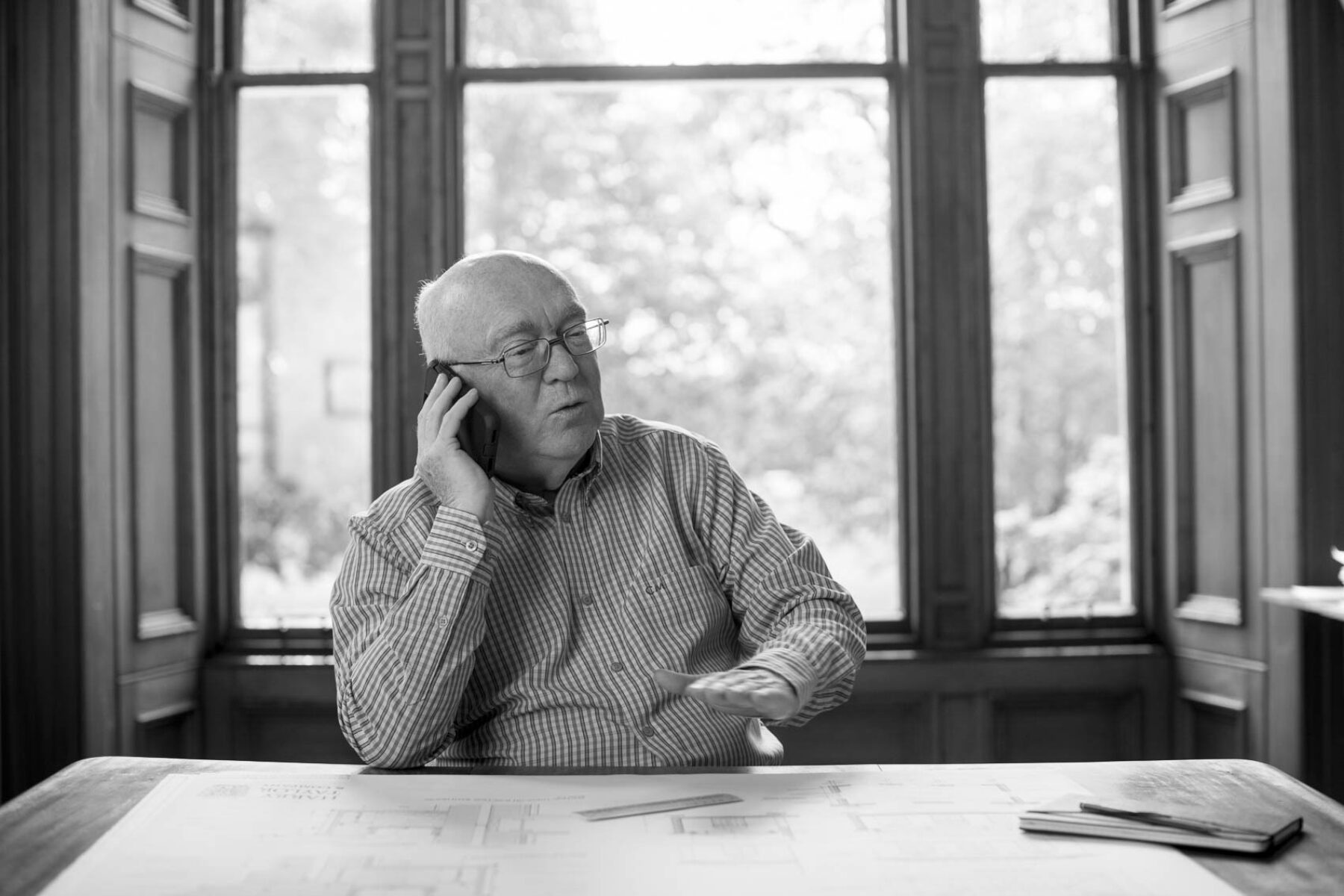 Black and white photo of Harry Taylor director speaking on the phone at his desk in front of a window.